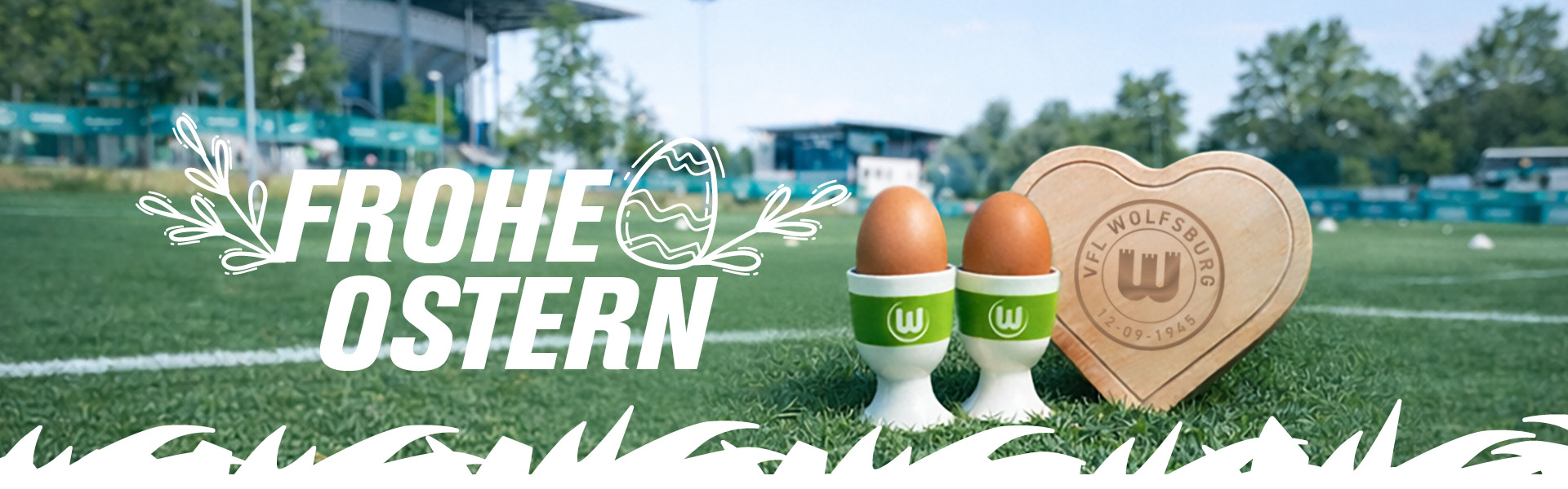 VfL Wolfsburg Ostergrafik mit „Frohe Ostern“ Schriftzug – zwei Eierbecher in Vereinsfarben mit Eiern und VfL-Logo sowie Holzherz mit Gravur auf dem Fußballplatz.