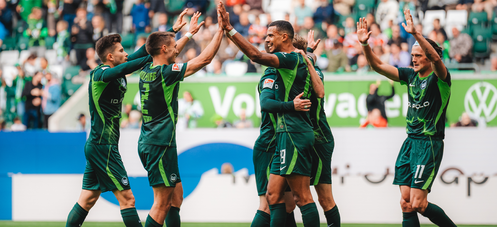 VfL-Wolfsburg-Spieler stehen auf dem Spielfeld beisammen und umarmen sich.