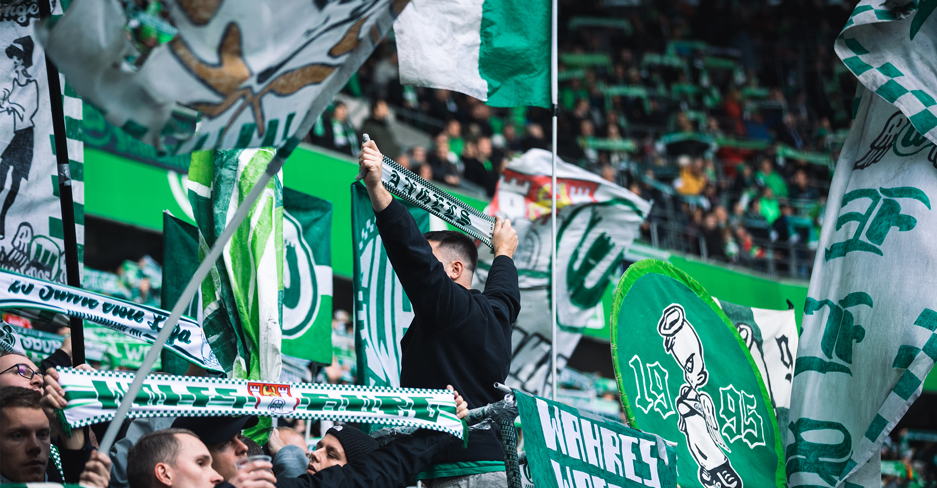 Eine Foto aus der Nordkurve, Fans halten Fahnen und Schals hoch und singen.