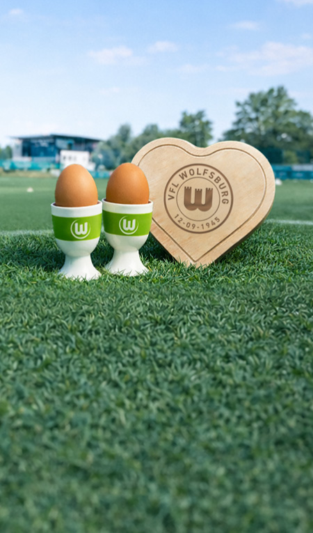 VfL Wolfsburg Eierbecher und Holzherz mit Logo auf dem Fußballrasen.