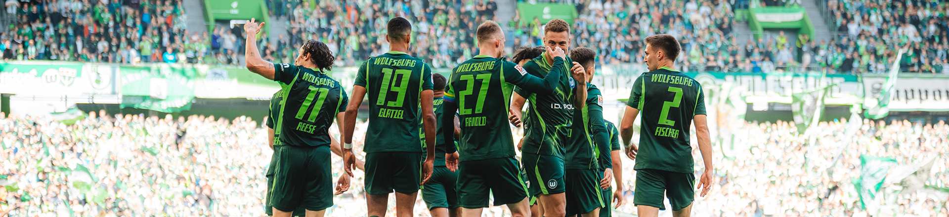 VfL-Wolfsburg-Spieler stehen vor VfL-Wolfsburg-Fans auf dem Spielfeld beisammen.