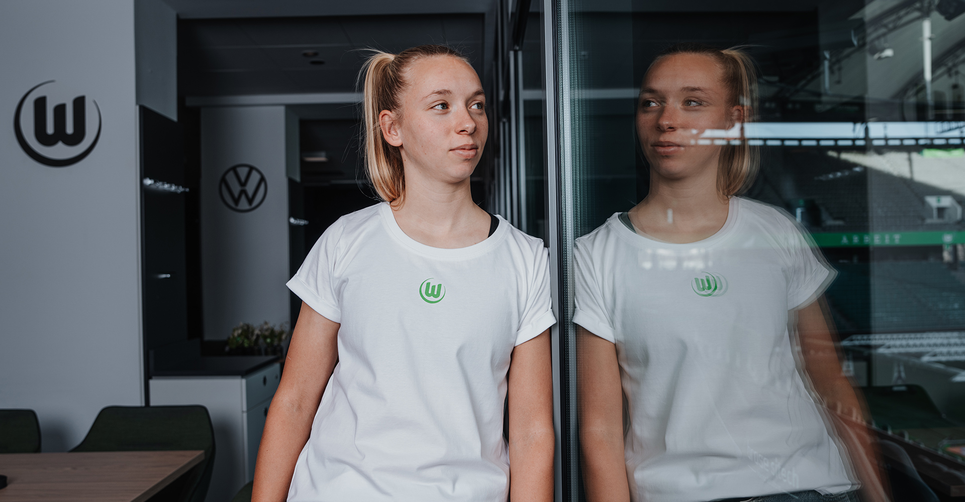 Eine VfL Wolfsburg Spielerin trägt ein weißes T-Shirt mit grünem VfL Wolfsburg Logo. Sie steht dabei am Fenster und guckt ins Stadion. Sie spiegelt sich dabei im Fenster.