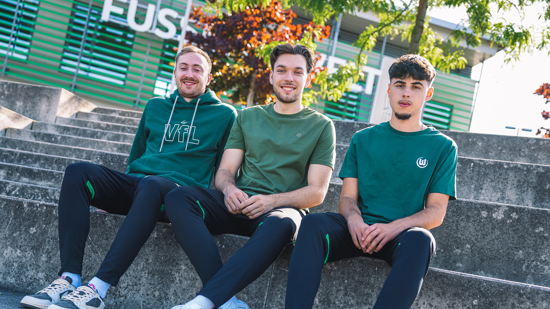 Drei VfL Fans sitzen auf der Treppe und tragen grüne T-Shirts und Hoodie vom vfL Wolfsburg