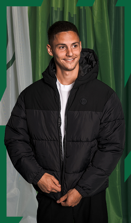 VfL-Wolfsburg-Spieler Bence Dardai trägt eine schwarze Steppjacke mit Kapuze und lächelt vor grün-weißem Hintergrund.